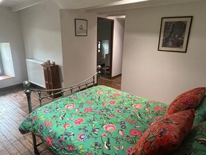 A bedroom with a floral bedspread, a wooden radiator, a metal bed frame, and a view of a hallway. - In addition to their room, our guests can enjoy a garden, a terrace, a veranda, and a lounge (Chevanceaux)