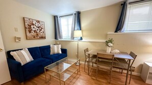 Een woonkamer met een blauwe bank, een glazen salontafel, een eettafel met stoelen, een lamp en een schilderij aan de muur.