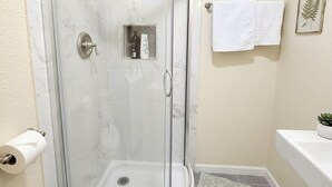Een douche met glazen wanden, een bedieningspaneel, een witte handdoek aan de muur en een kleine plant op de wastafel.