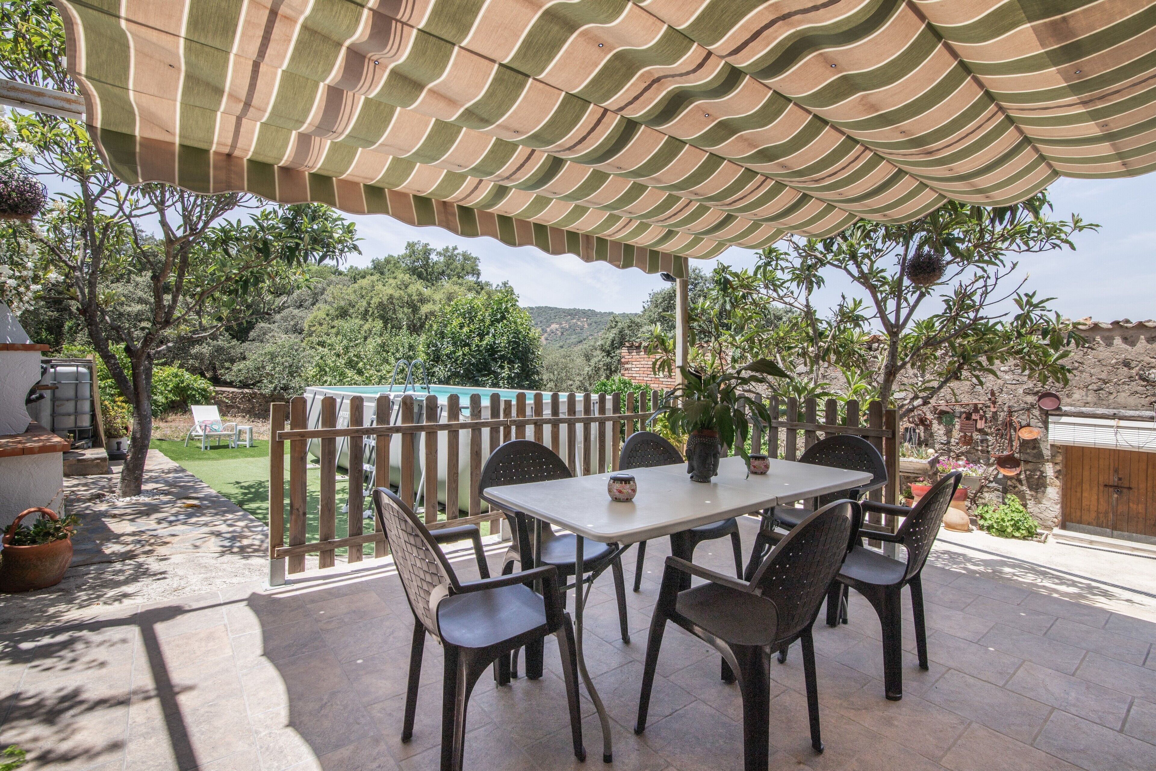 Een terras buiten met een gestreept zonnescherm, een tafel met stoelen, en uitzicht op een zwembad en groen.