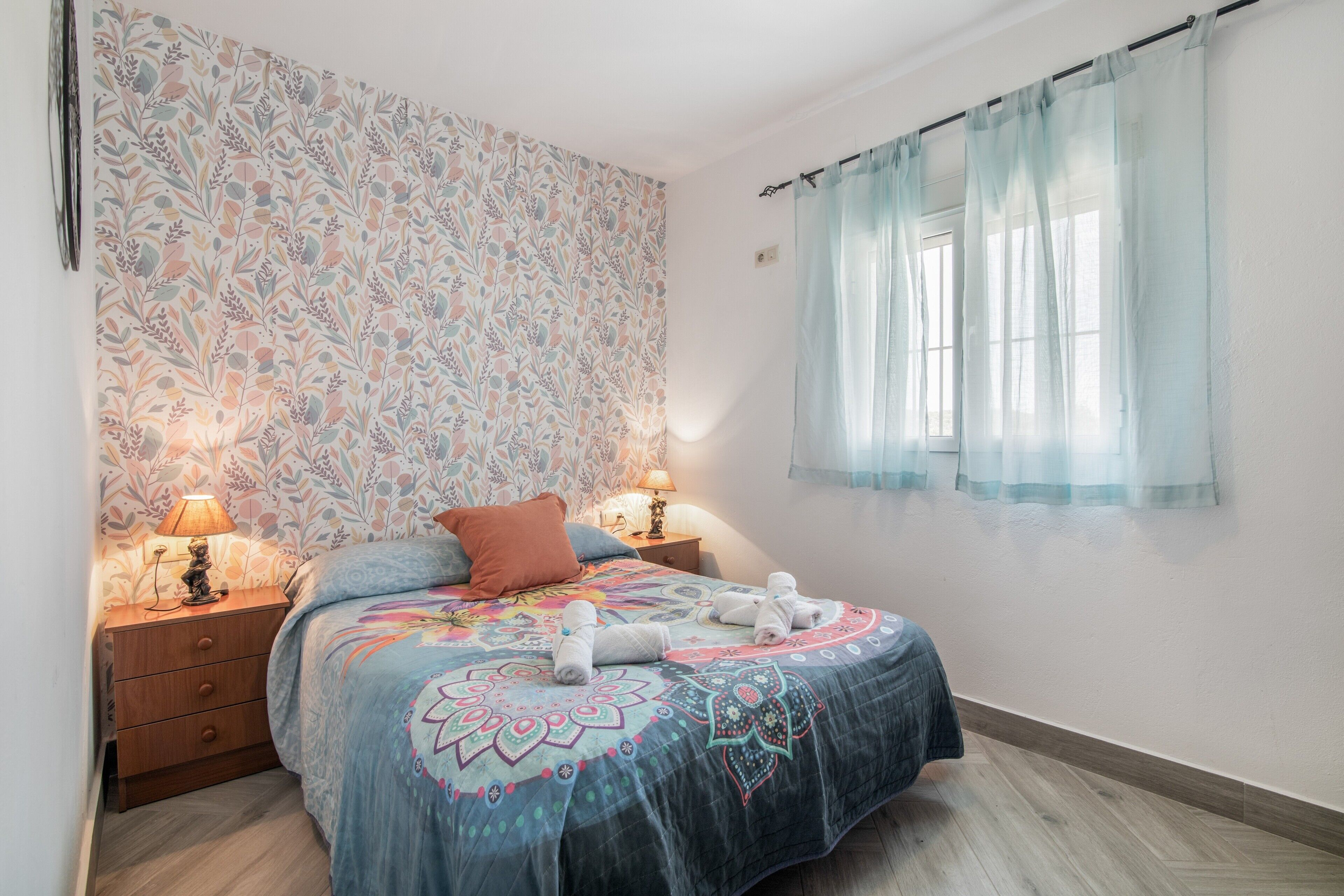 Une chambre à coucher avec un papier peint à motifs, un lit recouvert d’une couvre-lit à fleurs, deux lampes de chevet et une fenêtre ornée de rideaux légers.