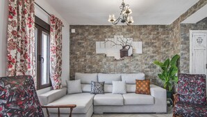 A living room with a sectional sofa, patterned armchairs, a chandelier, a potted plant, and a stone wall.