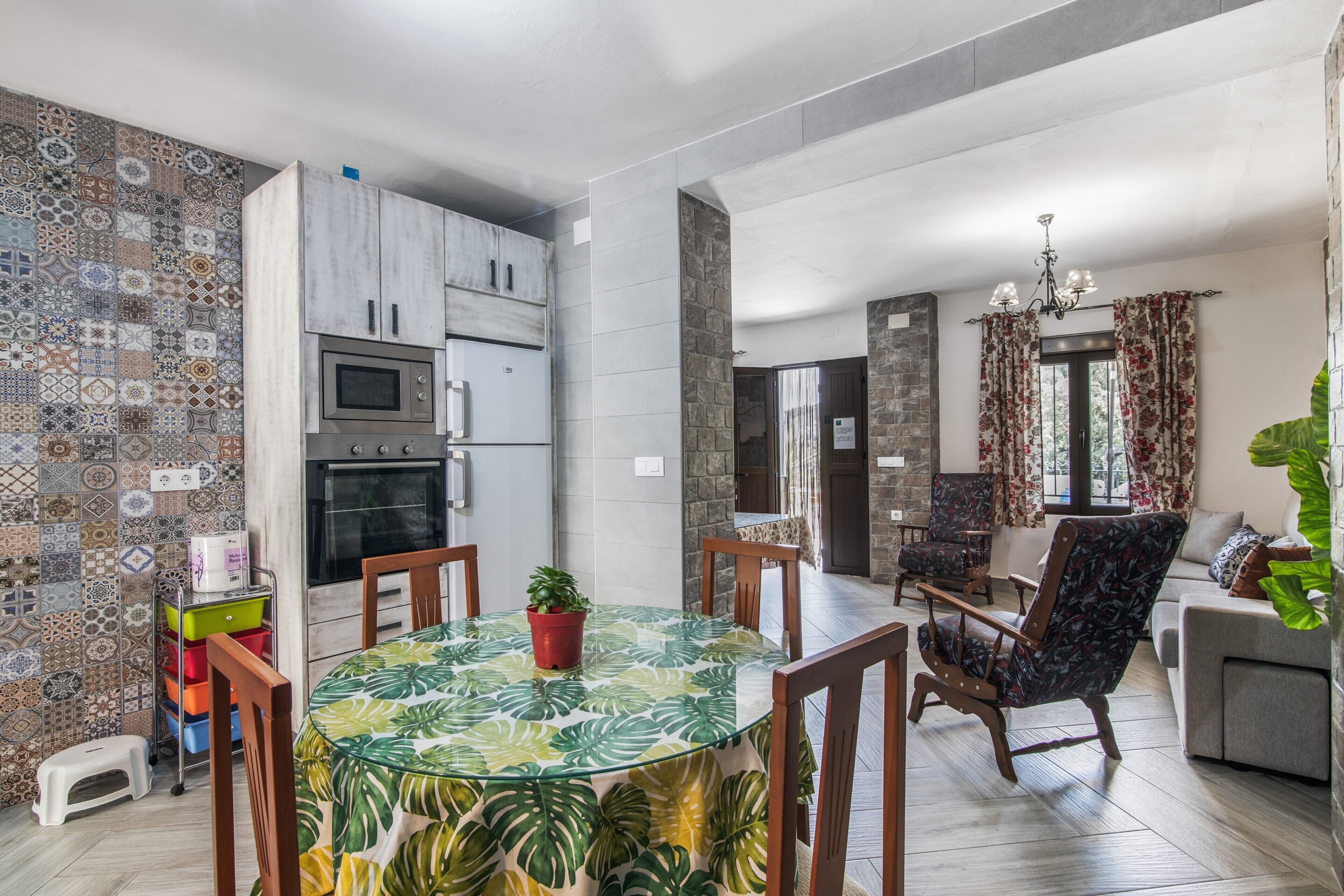 A modern kitchen with a microwave, oven, and cabinets, a dining area with a glass table and chairs, and a living room with a sofa and a large plant.