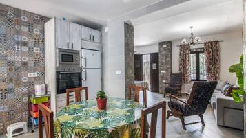 Una cocina moderna con microondas, horno y armarios, un comedor con mesa y sillas de vidrio, y una sala de estar con sofá y una planta grande.