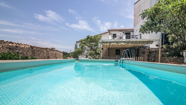 A swimming pool with a clear blue surface, surrounded by a stone wall and a building with a tiled roof.