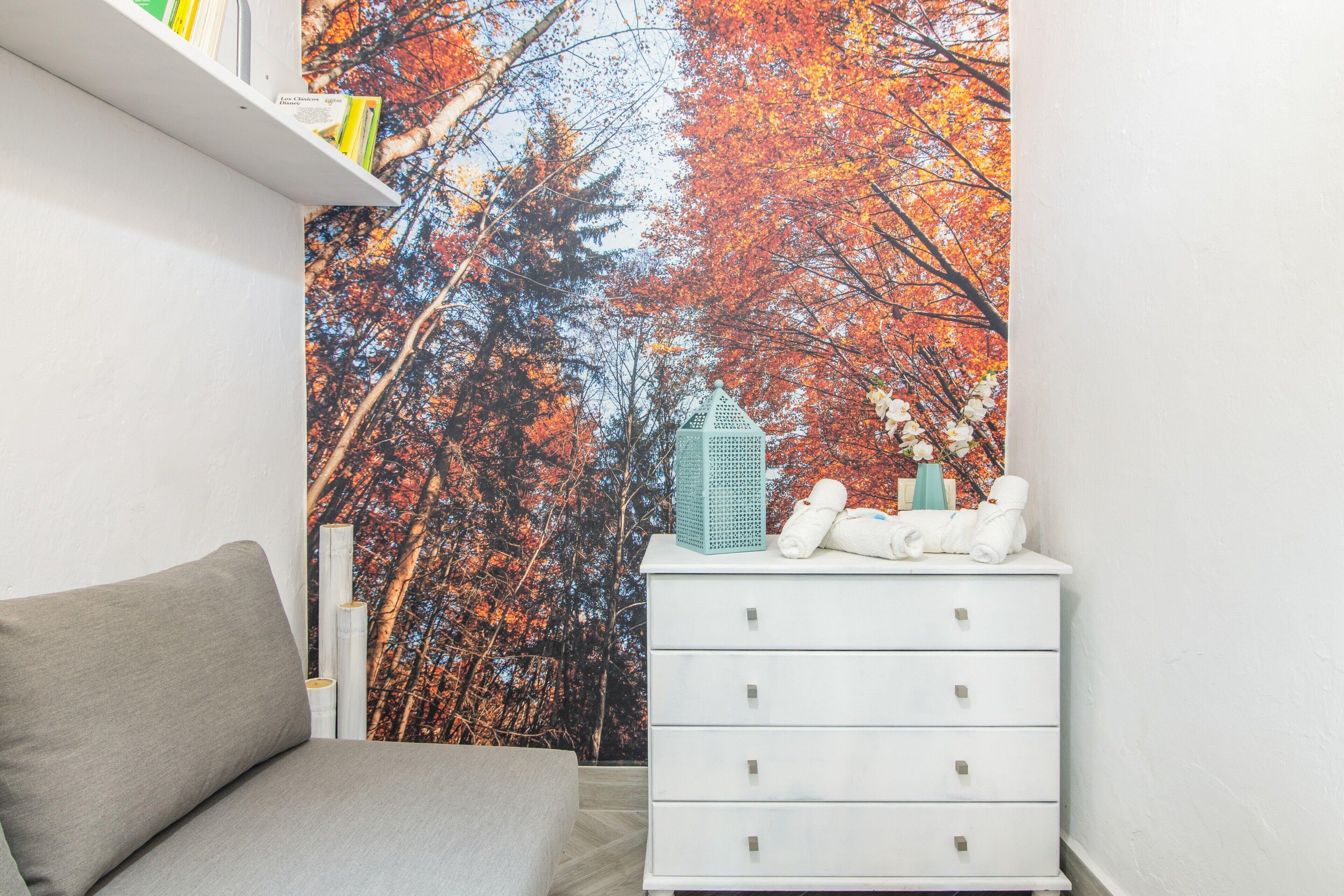 A bedroom with a bed, a white dresser, a shelf with books, and a wall mural of autumn trees.