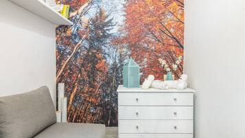 A bedroom with a bed, a white dresser, a shelf with books, and a wall mural of autumn trees.