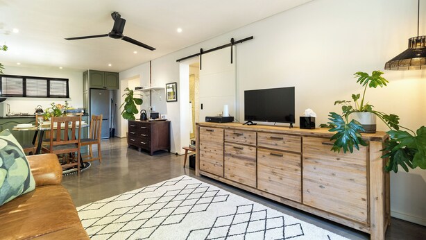 A modern kitchen with a dining area, wooden cabinets, a ceiling fan, and a TV on a wooden stand. - Scandi Garden Apartment (Stellenbosch)