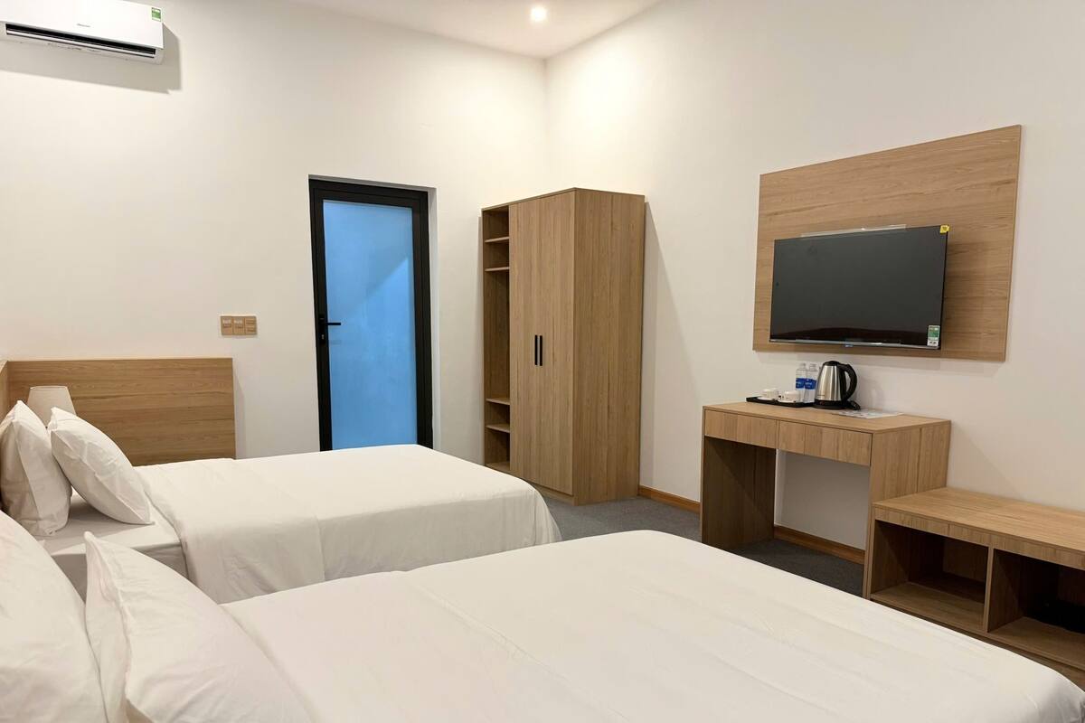 A hotel room with two beds, a desk with a TV, a wardrobe, and an air conditioning unit.