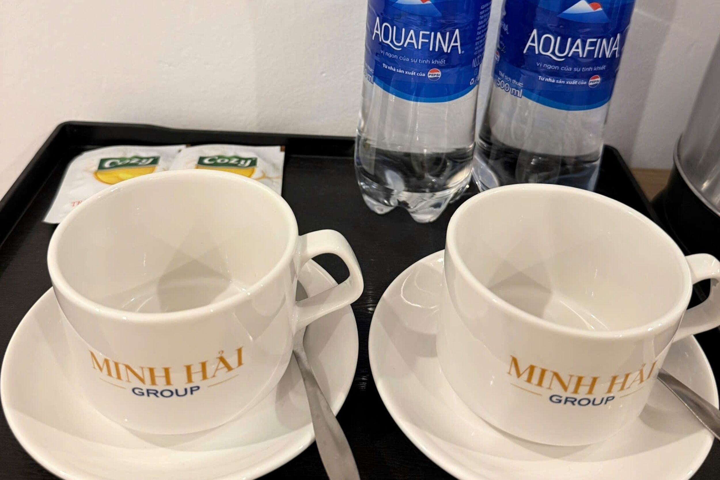 Two white cups with 'MINH HAI GROUP' text, a tray with two bottles of Aquafina water, and two packets of coffee.