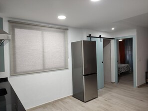 Une cuisine moderne dotée d’un réfrigérateur encastré, d’une fenêtre avec stores et d’un couloir menant à une chambre.
