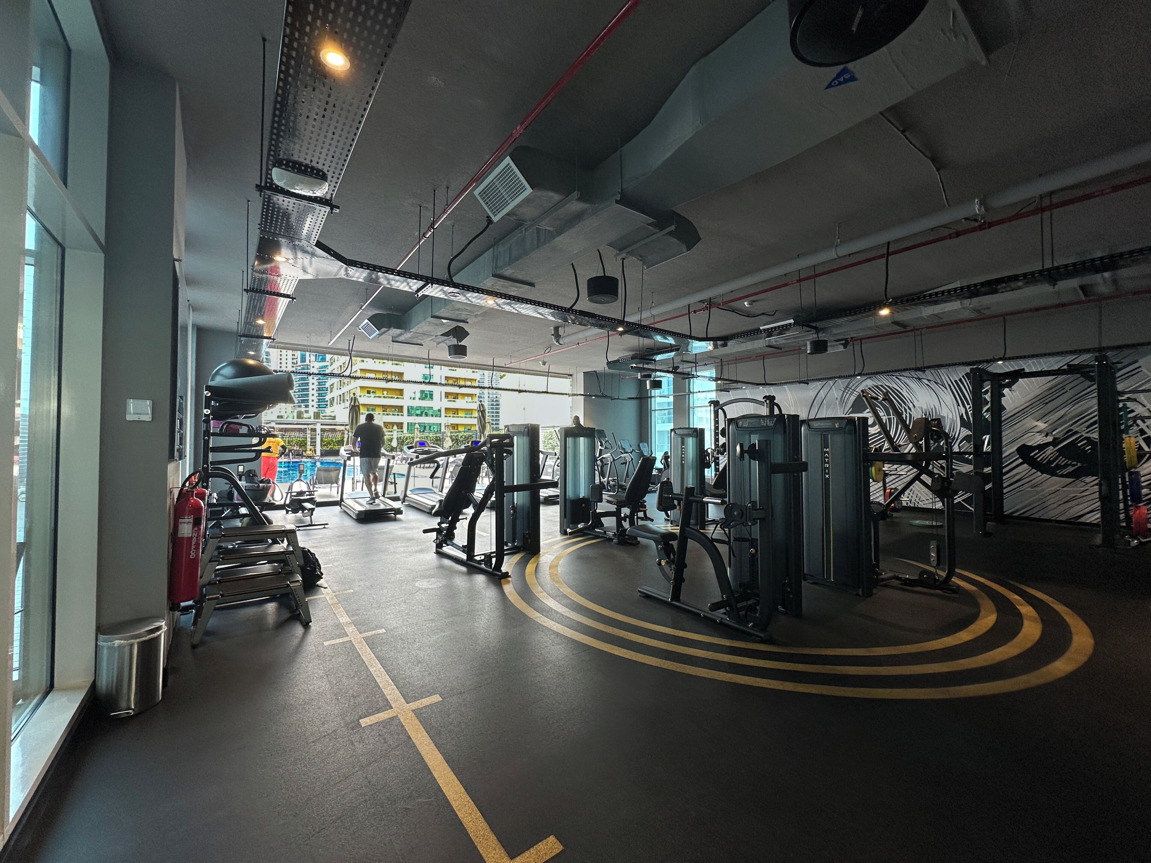 A gym with treadmills, weight machines, and a large window overlooking a building.