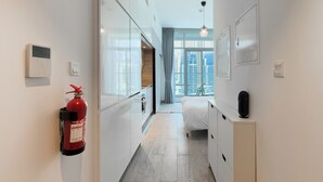En smal gang med en brandslukker ophængt på væggen, en seng med hvidt sengegavl og et lille skab med en sort genstand ovenpå.