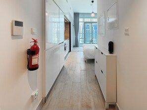 A narrow hallway with a fire extinguisher mounted on the wall, a bed with a white headboard, and a small cabinet with a black object on top. - Modern Studio in Prime Location & JBR (Dubai Marina)