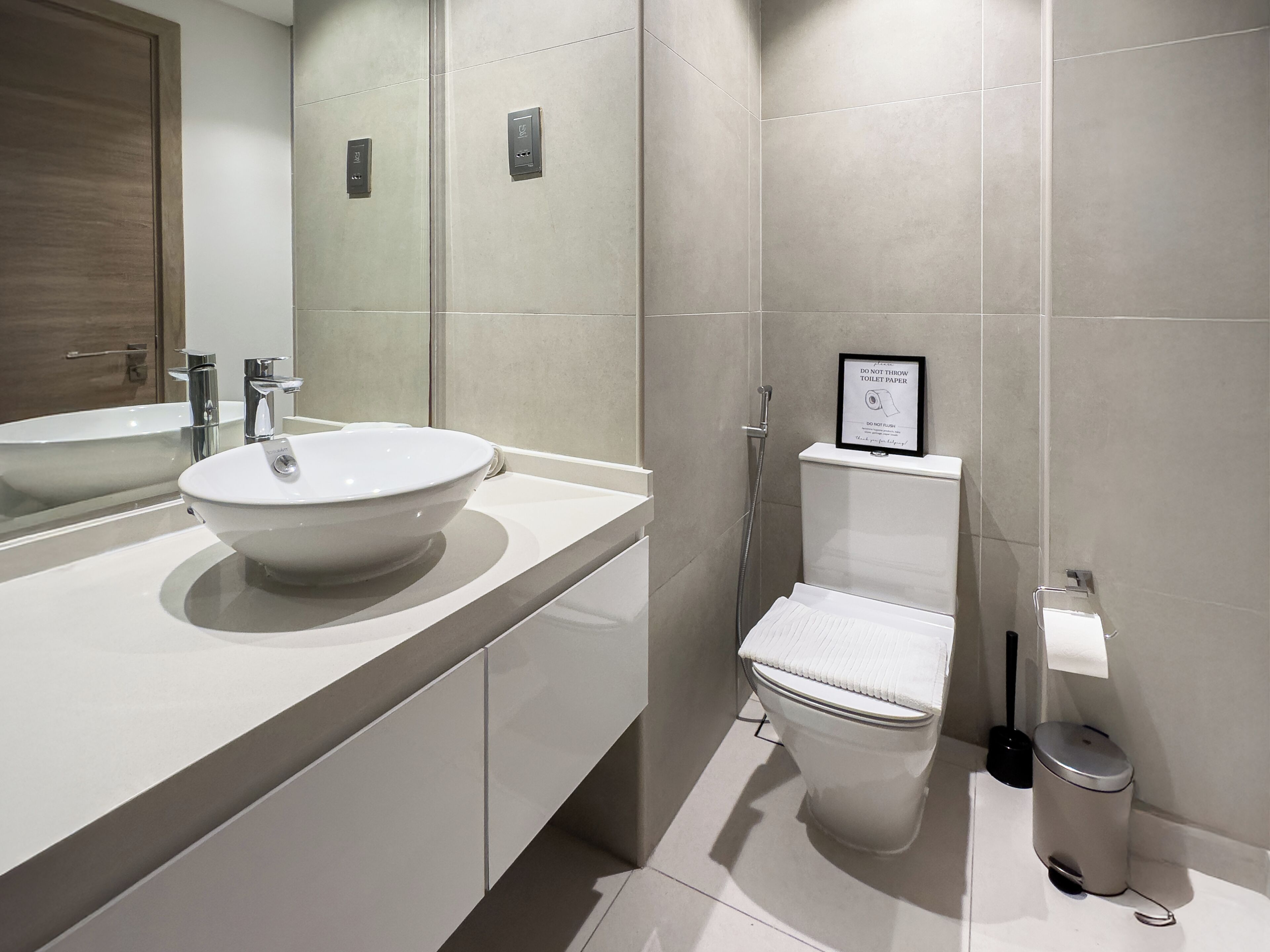 Een moderne badkamer met een grote spiegel, een wit wastafelmeubel, een toilet en een prullenbak.