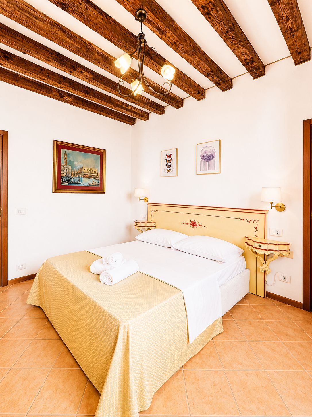A hotel room with a bed, wooden ceiling beams, a painting, two bedside lamps, and a bedside table.