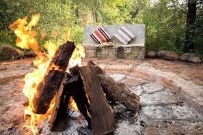 Ein Lagerfeuer mit Holzscheiten, einer Steinfürstelle und zwei gestreiften Kissen auf einer Bank.