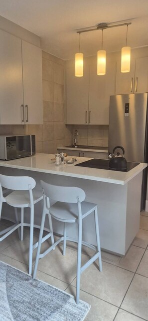 Eine moderne Küche mit einer zentralen Kochinsel, weißen Barhocker, Edelstahlgeräten und Pendelleuchten.