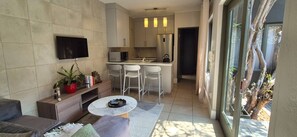 A modern living room with a sofa, a coffee table, and a TV mounted on the wall. There is a kitchen area with a refrigerator, microwave, and bar stools. - Cozy 2 Bedroom Cottage (Johannesburg)