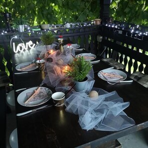 An outdoor dining area with tables set for two, featuring a neon sign, string lights, and potted plants. - Holiday house Malia Spa Hot Tub Jacuzzijem Free entry to all City attractions (Čepelovac)