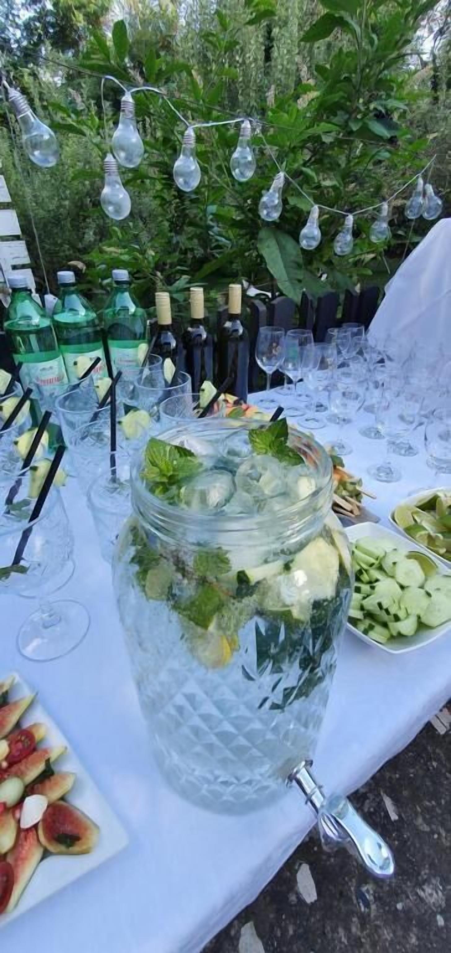 A table with a large mason jar filled with a beverage, surrounded by glasses and bottles, with a plate of fruit and a white tablecloth.