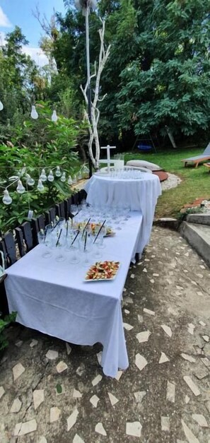 An outdoor dining area with tables covered in white tablecloths, surrounded by greenery and a stone-tiled floor.