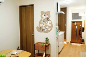 Ein Zimmer mit Holztür, einem an der Wand befestigten Schwarzdbrett, einem kleinen Tisch mit Tasse sowie einer Küchenzeile mit weißem Spülbecken und Schränken.