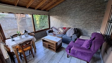 A cozy room with a wooden ceiling, stone wall, a purple sofa, a blue sofa, a wooden table with chairs, and a window with a view of greenery.