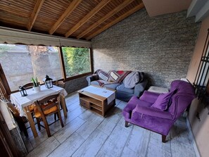 Una habitación acogedora con techo de madera, pared de piedra, un sofá púrpura, un sofá azul, una mesa de madera con sillas y una ventana con vistas a la vegetación.