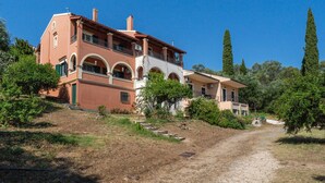 A two-story house with a red facade, multiple balconies, and a dirt path leading to the entrance. - Apartment 'Villa Boho' with Sea View and Shared Garden (Kato Korakiana)