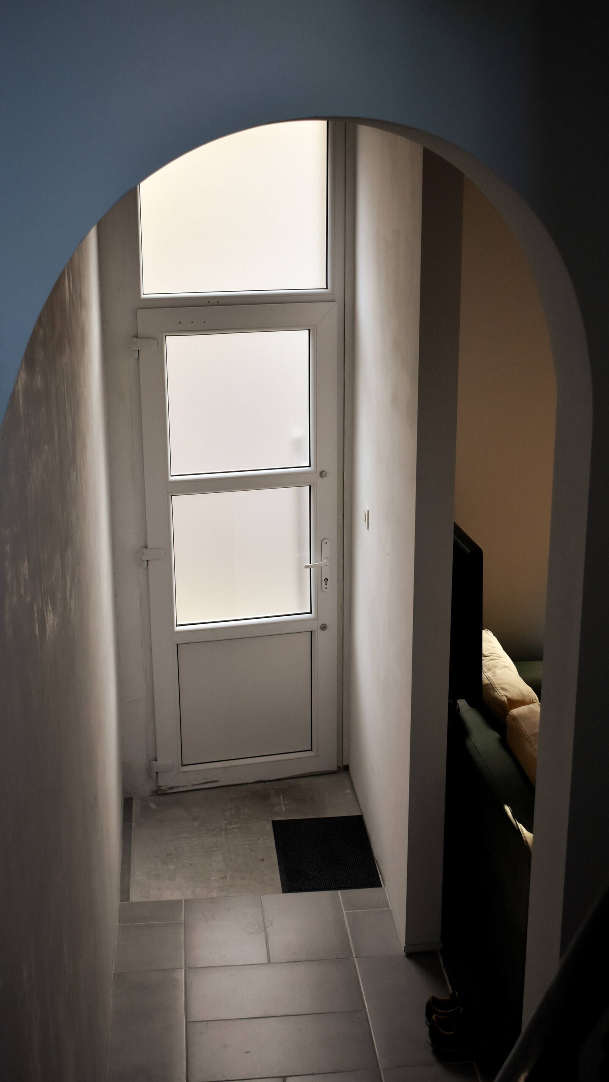 Un vestíbul estret que condueix a una porta amb una finestra que ofereix vistes, un estri de peu negre a la base i un sofà de color fosc a l'habitació adjacenta.