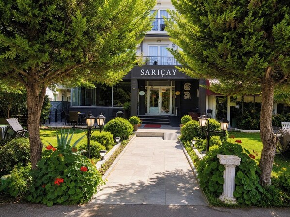 Un edificio con un letrero que dice 'SARICAY' en la entrada, rodeado de follaje y mobiliario exterior.