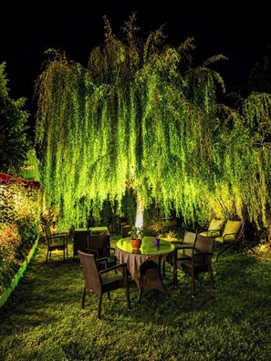 Un área de comedor al aire libre con una mesa y sillas, rodeada de exuberante vegetación y un sauce llorón.