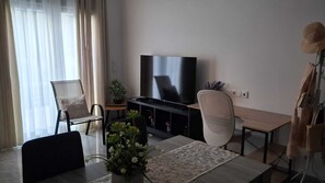 Una sala de estar con televisor, una mesa de comedor, sillas, un sofá, una planta en maceta y un perchero.