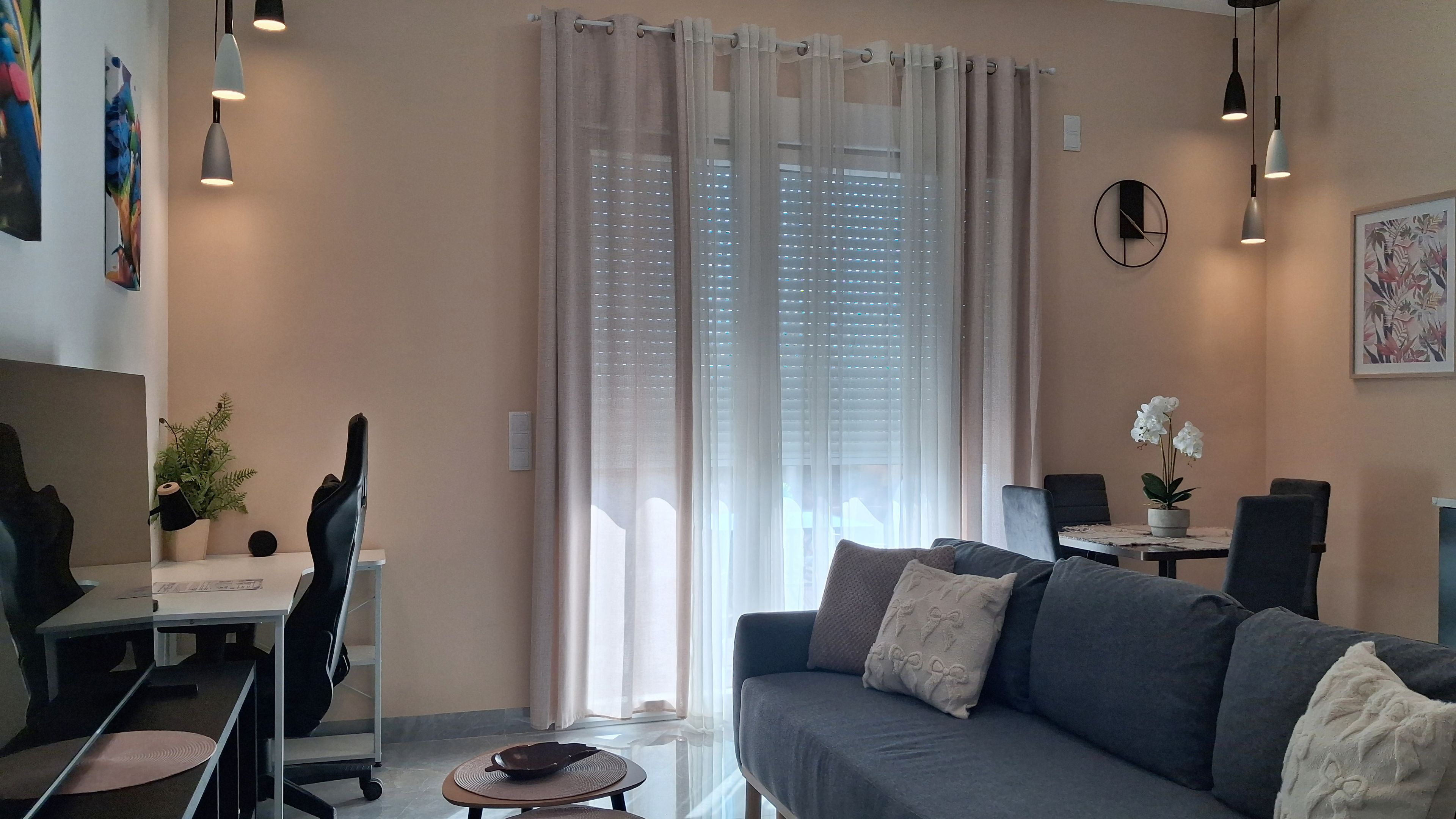 A modern living room with a grey sofa, a dining table with chairs, a desk with a chair, and a large window with sheer curtains.