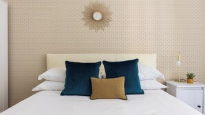 Un lit bien fait, agrémenté d’oreillers blancs et turquoise, un élément décoratif doré accroché au mur et une table de chevet avec une lampe et une petite plante.