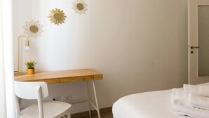 Une chambre avec un bureau en bois, une chaise blanche, un lit avec une literie blanche et trois œuvres d’art accrochées au mur.