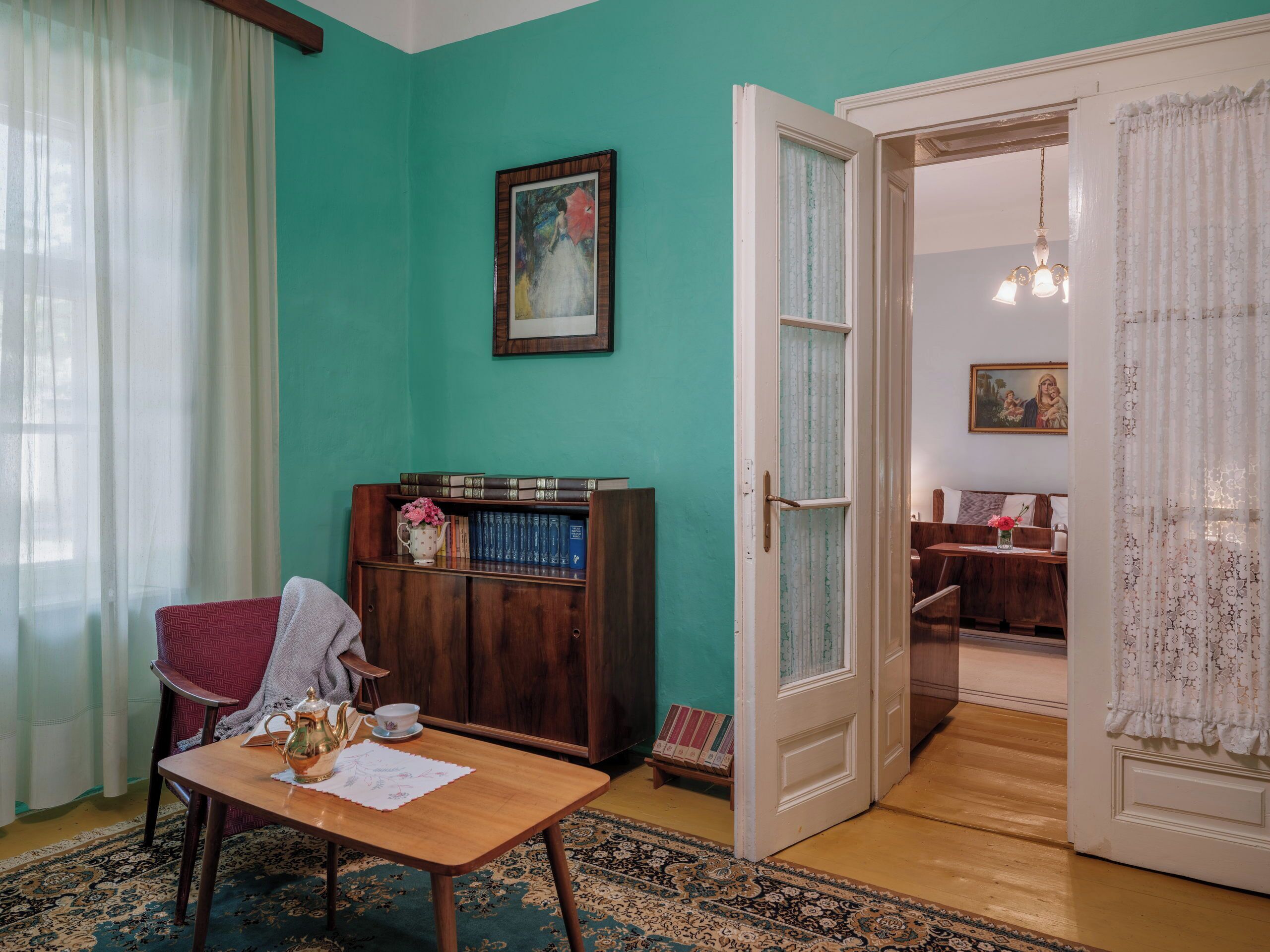 Ein Zimmer mit türkisfarbenen Wänden, einem Holztisch mit Teekanne und Tasse, einem Bücherregal und einer Tür zu einem anderen Raum.
