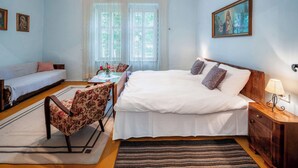 Een slaapkamer met een bed, een slaapbank, een eettafel met stoelen, een kroonluchter en een schilderij aan de muur.