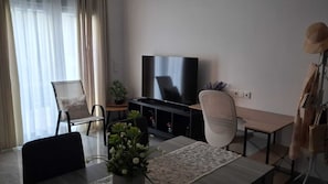 A living room with a TV, a dining table, chairs, a sofa, a potted plant, and a coat rack.