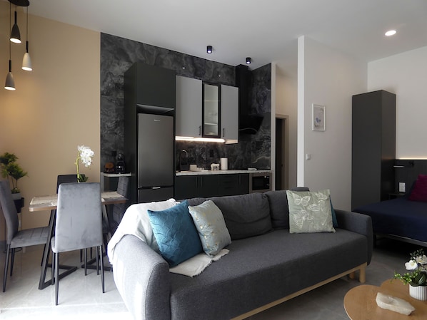 A modern living room with a grey sofa, a dining area with a table and chairs, a kitchen with dark cabinetry, and a small side table with a plant.