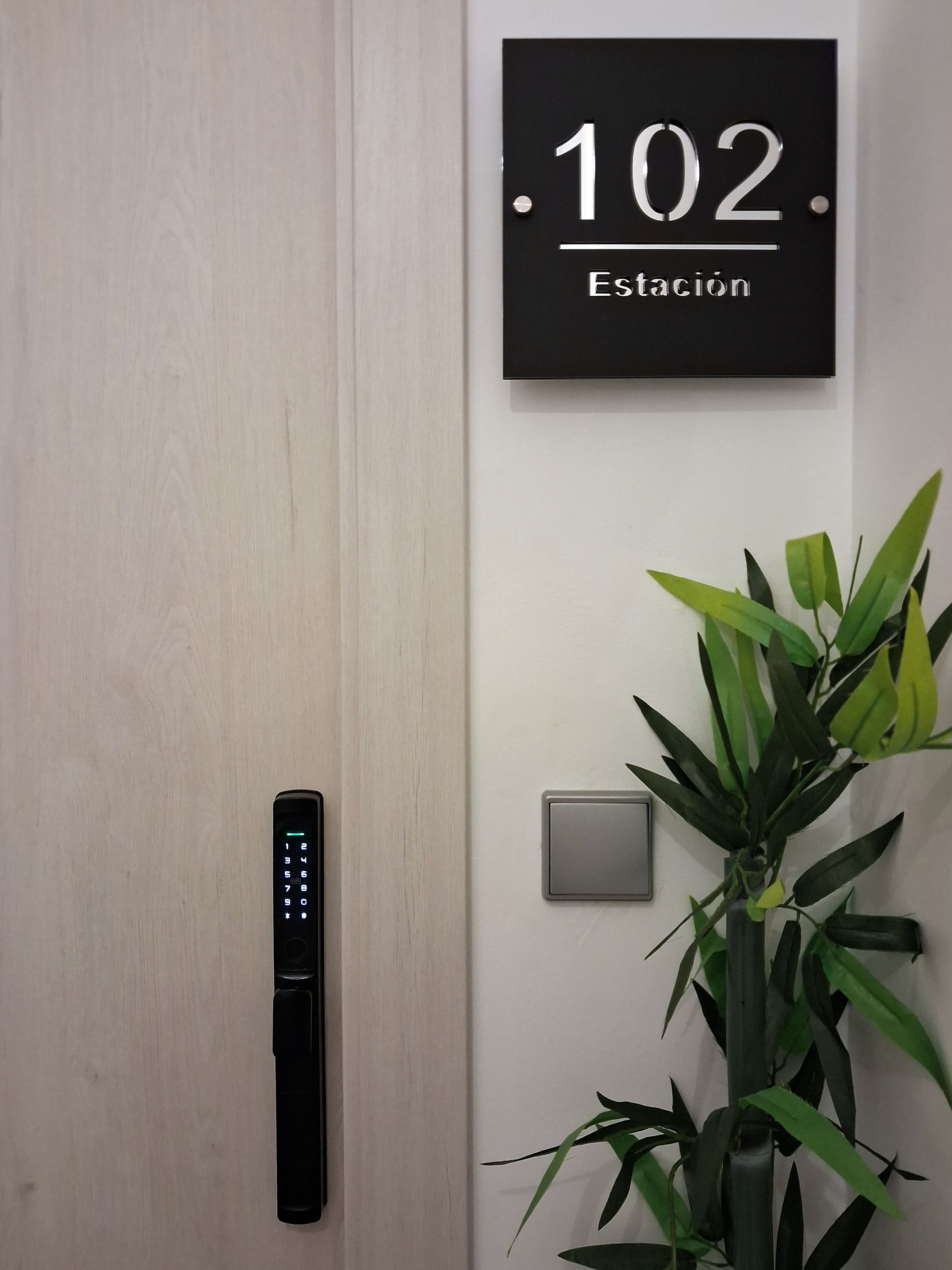 Una puerta con cerradura electrónica de teclado numérico, un cartel con el número de habitación 102 y una planta en maceta junto a la pared.