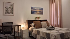 Une chambre d’hôtel avec un lit, un bureau, une chaise, une table de chevet, une lampe, un tableau et une fenêtre avec des rideaux.