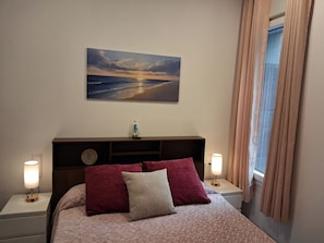 Un dormitori amb un llit, una tauleta de nit amb una làmpada, un quadre penjat a la paret i una finestra amb cortines.