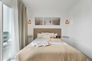 A hotel room with a bed, a window with curtains, a bedside table with a tray, and wall art. - Charming Apartment in Arona Comfort and Convenience Await (Arona)