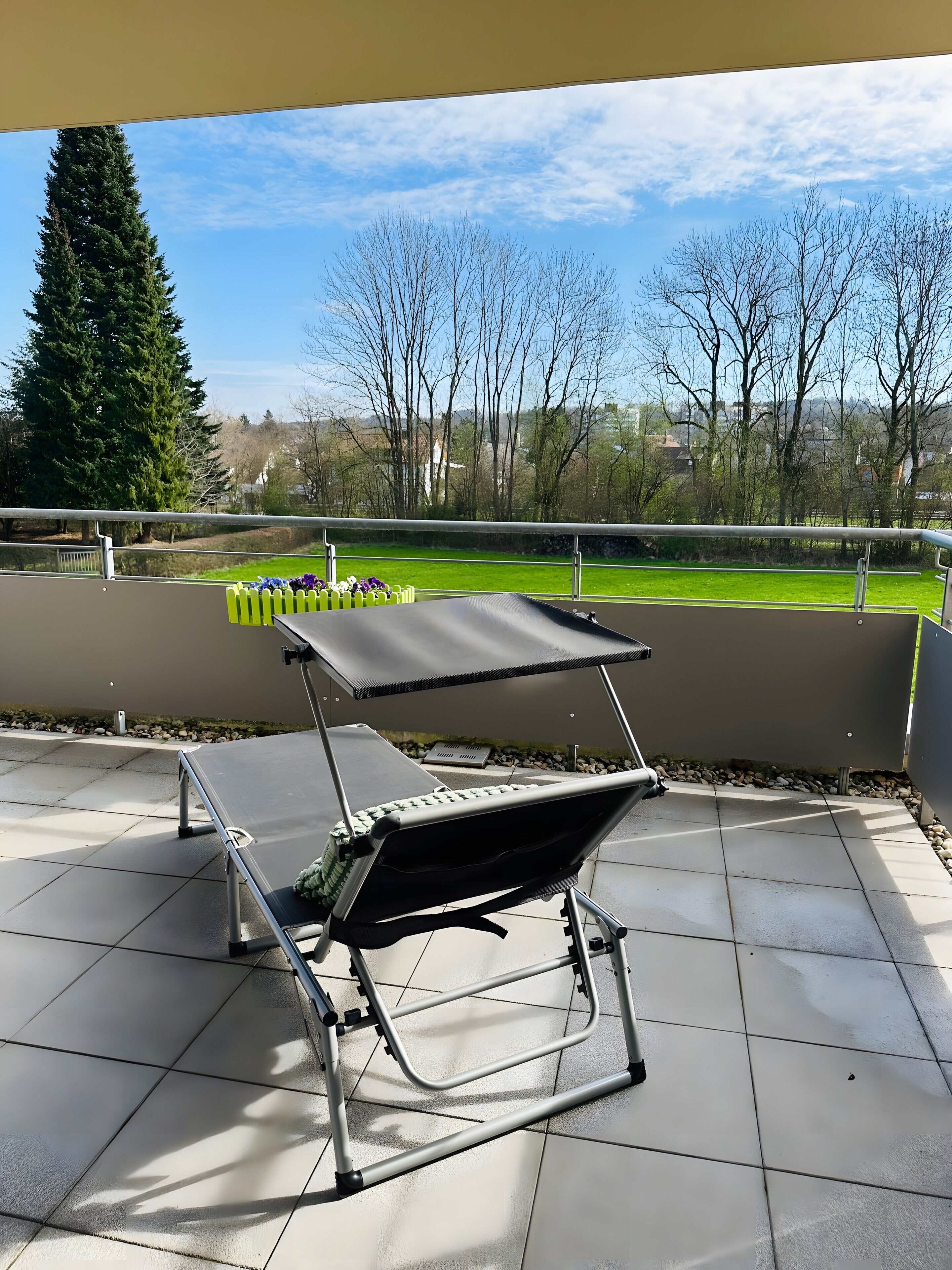 Eine Terrasse mit Klapptisch und -stühlen, Blick auf einen Park mit Bäumen und einer Rasenfläche.