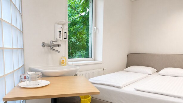 Habitación de hotel con cama, una mesa con silla, ventana, lavamanos y cubo de basura.