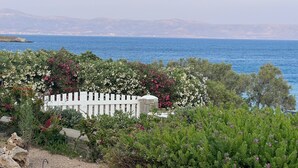 A coastal landscape with a white fence, dense greenery, and a view of the sea and distant mountains. - Picturesque home with amazing view (Paros)