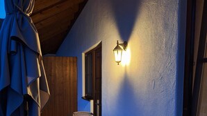 Ein überdachter Außenbereich mit runden Tischen und Korbstühlen, einer Holzbalkendecke und einer an der Wand befestigten Leuchte.