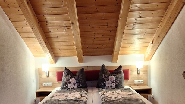 Ein Doppelbett mit floralem Bettzeug in einem Zimmer mit Holzbalken und schräg versiegelter Decke.